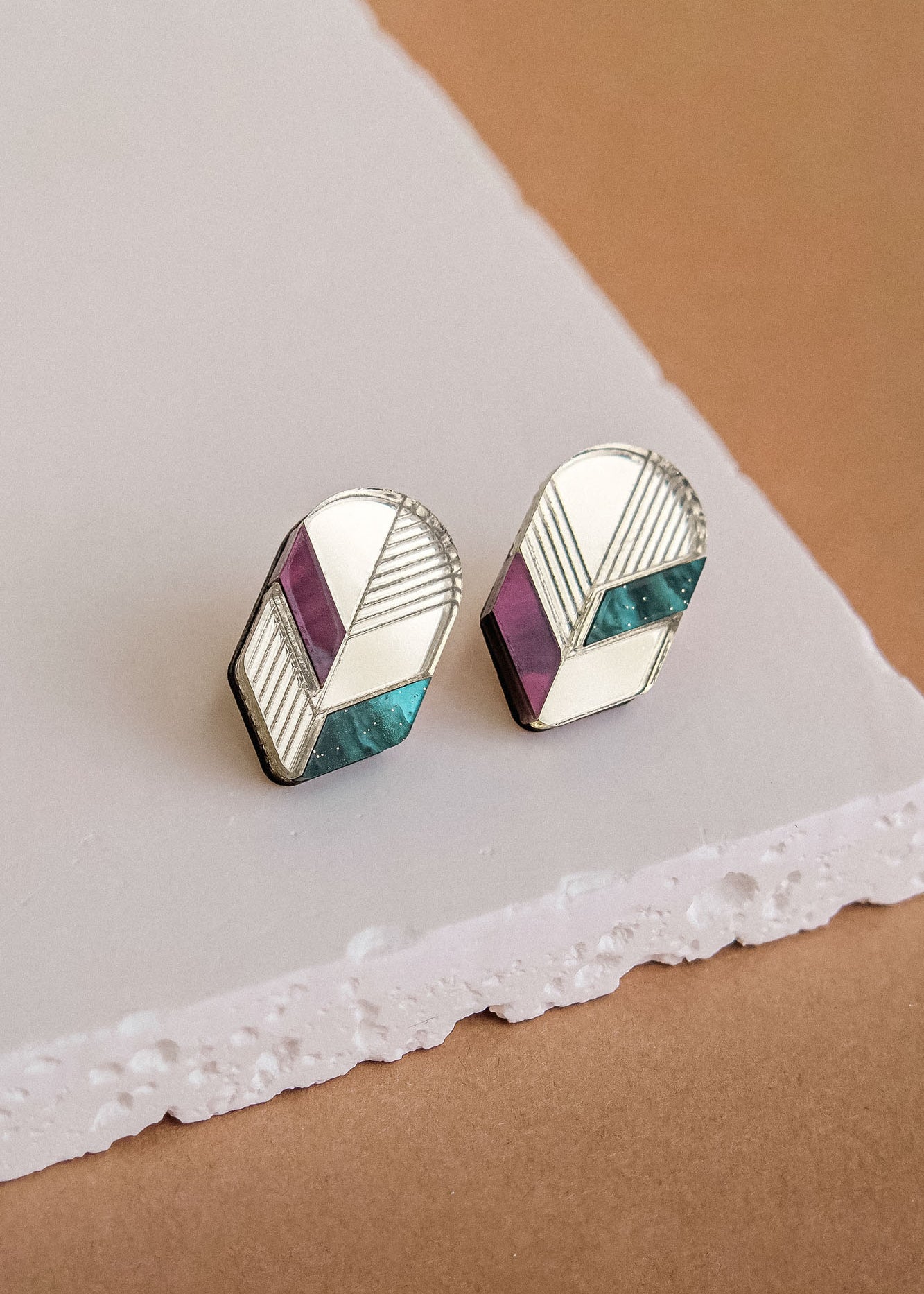A pair of geometric stud earrings with silver outlines, featuring sections of teal, purple, and cream enamel, perfect for lovers of minimalist jewelry and displayed on a textured white and brown surface.
