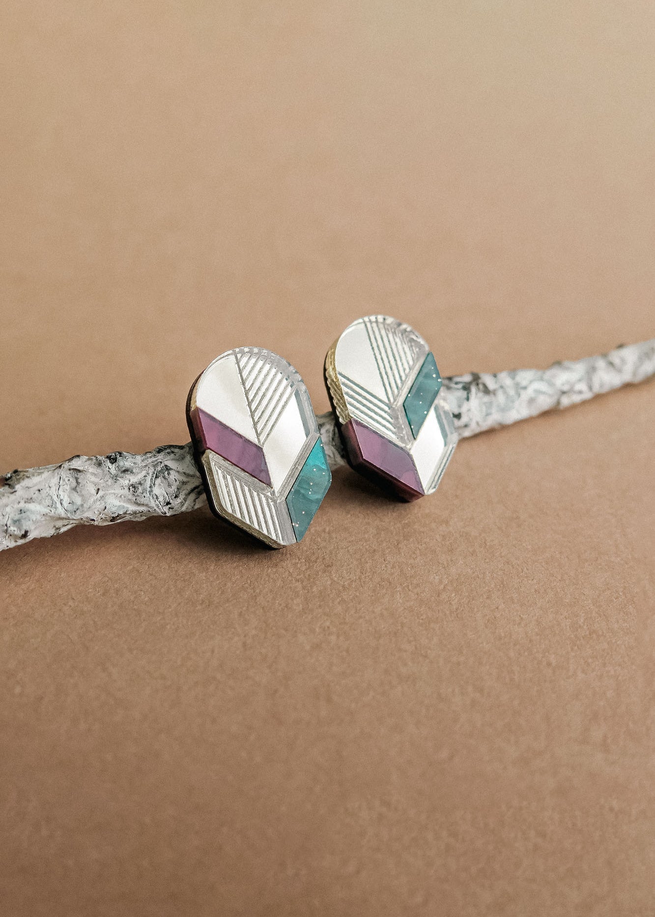 Handmade geometric stud earrings with silver, lavender, and teal sections rest on a textured, light gray branch against a beige background—a perfect touch of minimalist jewelry.