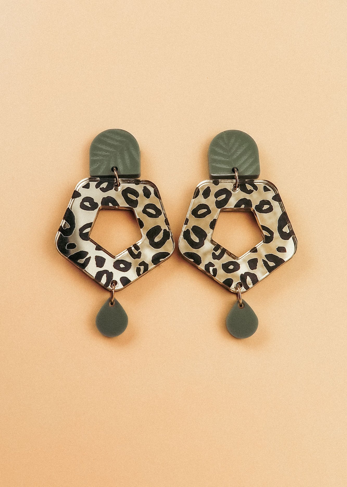 A pair of Leopard Earrings with olive green tops and teardrop-shaped dangles, featuring hexagonal centers in timeless patterns, crafted from premium materials and displayed on a peach background.
