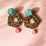 A pair of Leopard Earrings featuring green leaf-shaped tops, hexagonal leopard-print centers with cutouts, and small pink teardrop dangles—displayed on a light pink background for standout style with timeless patterns.