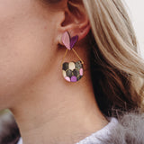 Close-up of a woman's ear wearing playful Mermaid Tail Earrings with a pink and purple heart-shaped stud and a semi-circular dangle in green, pink, and purple—a perfect accessory for mermaid lovers. The woman has blonde hair and wears a white top.