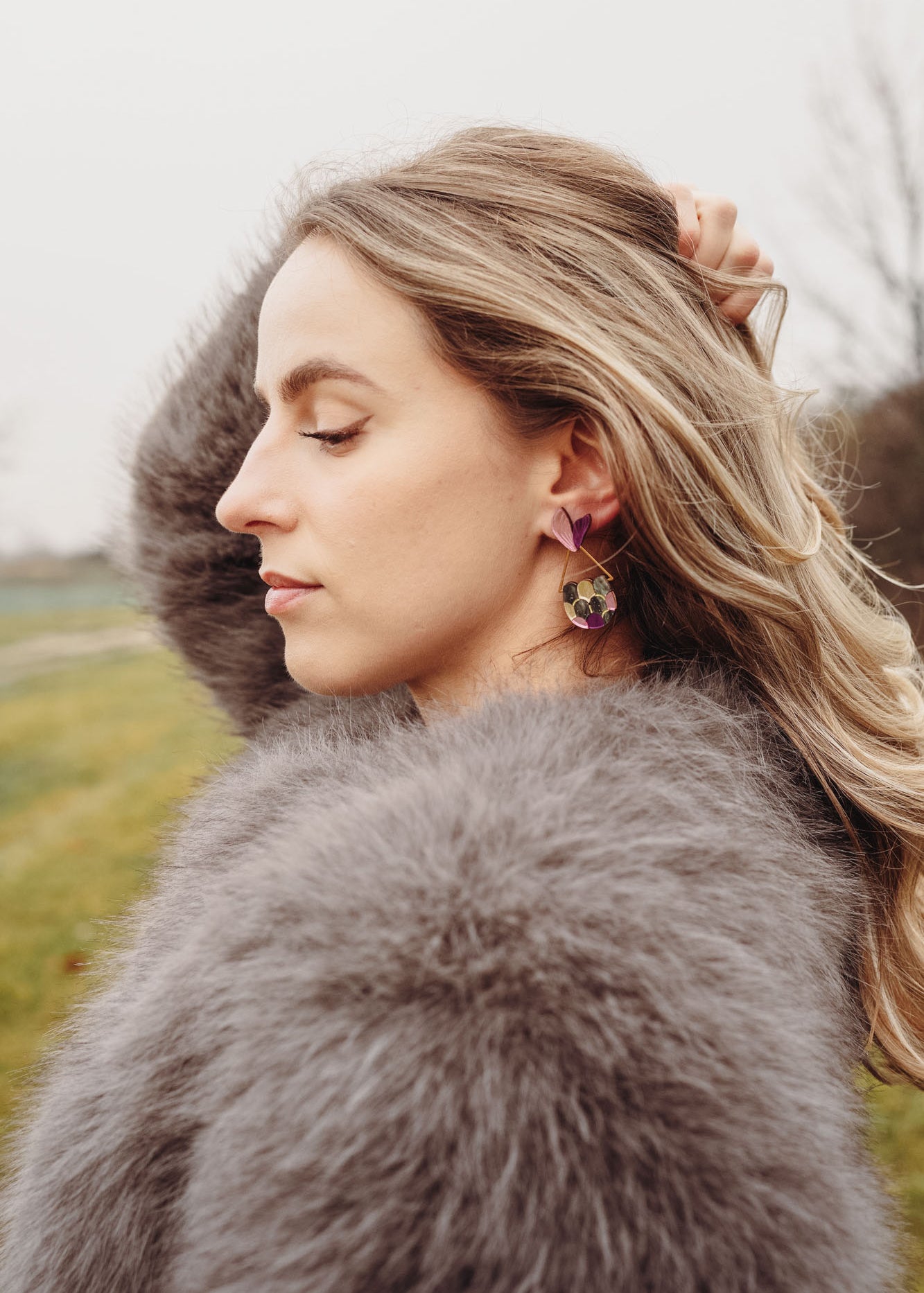 A woman with wavy blonde hair, wearing a fluffy gray coat and playful Mermaid Tail Earrings, stands outdoors with her eyes closed and one hand in her hair. The background is grassy with blurred trees—perfect for mermaid lovers.