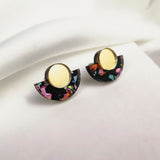 Minimalist Geometric Stud Earrings featuring gold circular tops and black semicircular bottoms with colorful, confetti-like patterns. These lightweight, hypoallergenic earrings are displayed on a white fabric background.