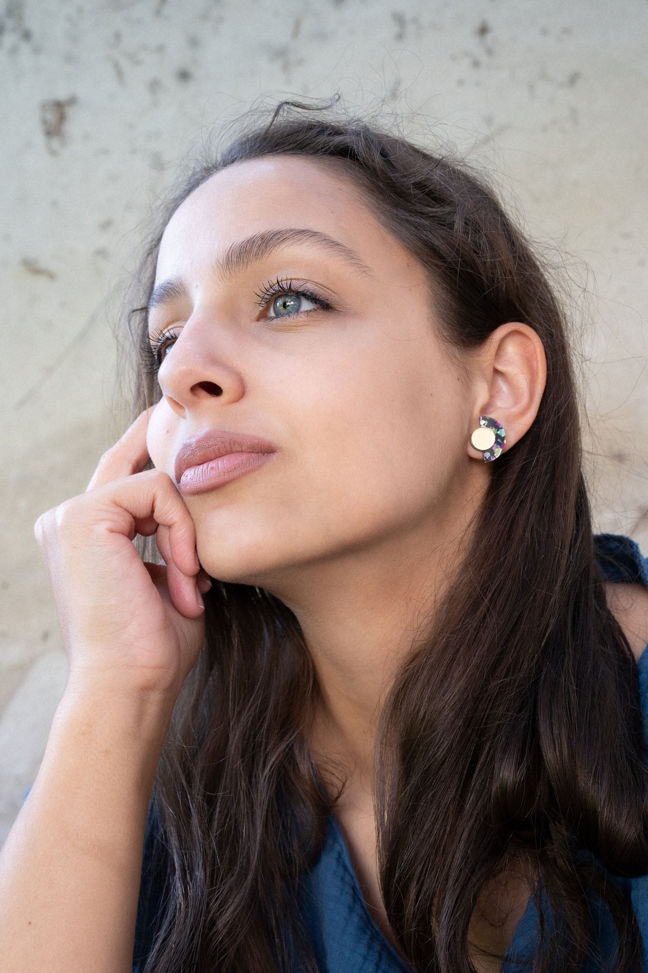 A woman with long brown hair and light eyes looks thoughtfully into the distance, resting her chin on her hand. She is wearing a blue top and minimalist geometric stud earrings, with a neutral, textured background behind her.