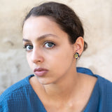 A young woman with light eyes and dark hair pulled back, wearing a textured blue top and hypoallergenic Minimalist Geometric Stud Earrings, gazes seriously at the camera against a neutral, slightly textured background.