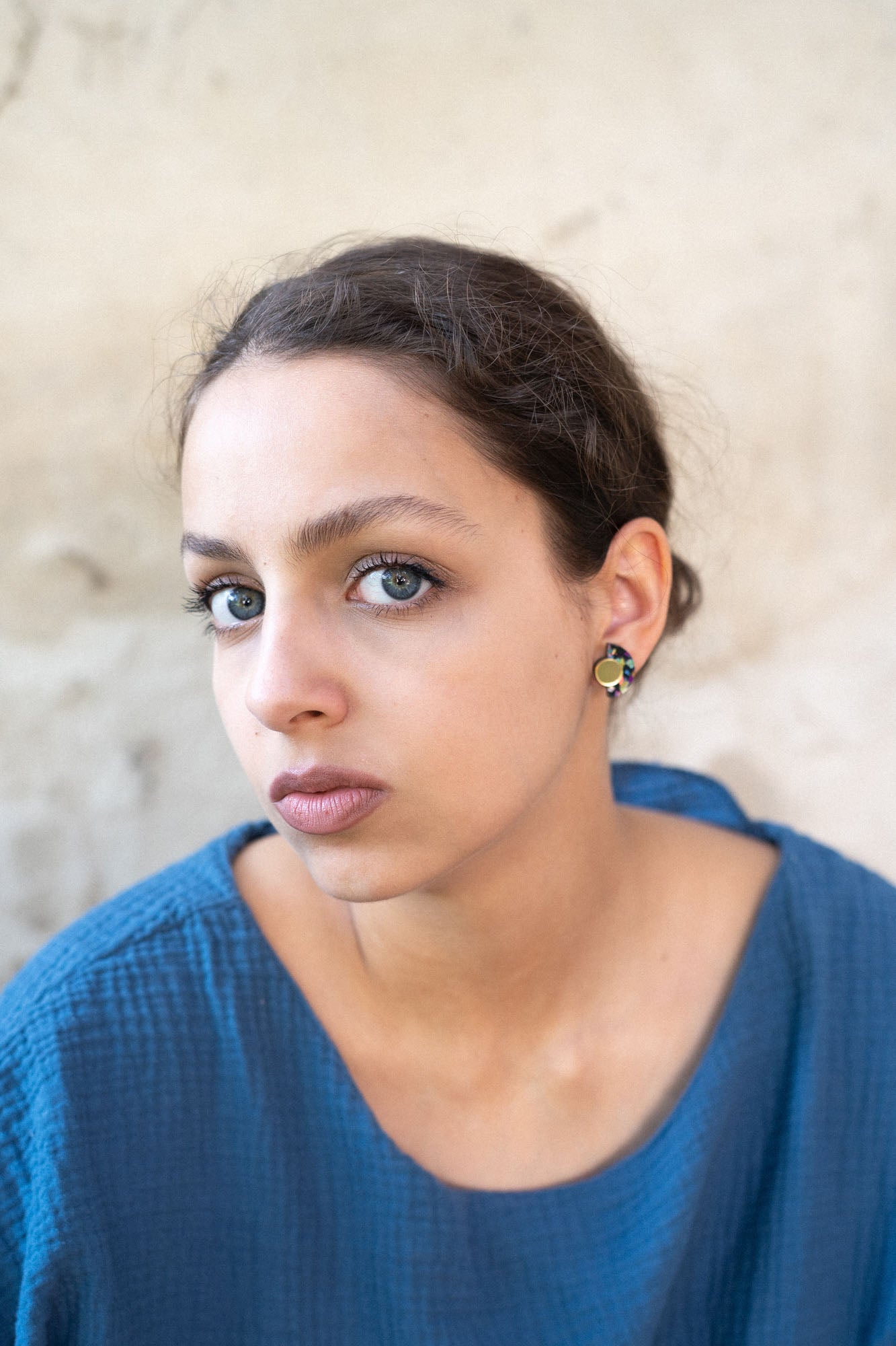 A young woman with light eyes and dark hair pulled back, wearing a textured blue top and hypoallergenic Minimalist Geometric Stud Earrings, gazes seriously at the camera against a neutral, slightly textured background.