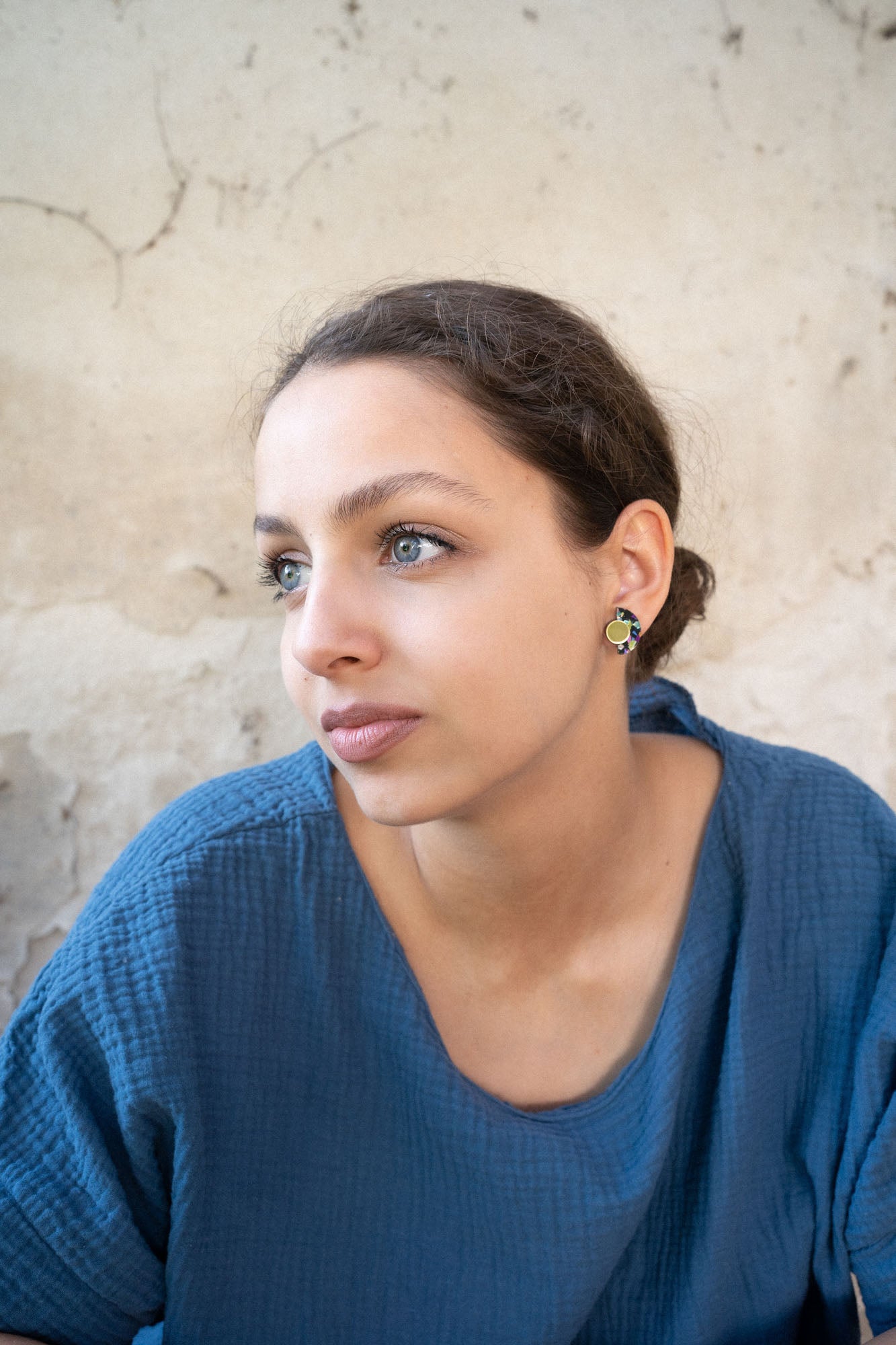 A young woman with light eyes and brown hair pulled back, wearing a textured blue top and lightweight Minimalist Geometric Stud Earrings, looks thoughtfully to the side against a beige, textured wall.