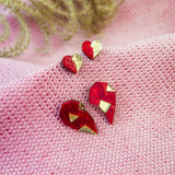 Red and gold geometric heart studs and pins, perfect as romantic jewelry, are displayed on textured pink fabric, with decorative plant stems in the background.