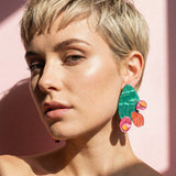 A woman with short blonde hair and glowy skin poses in sunlight, wearing bold design statement earrings shaped like opuncia cactus with green, pink, and orange details, against a pink background.