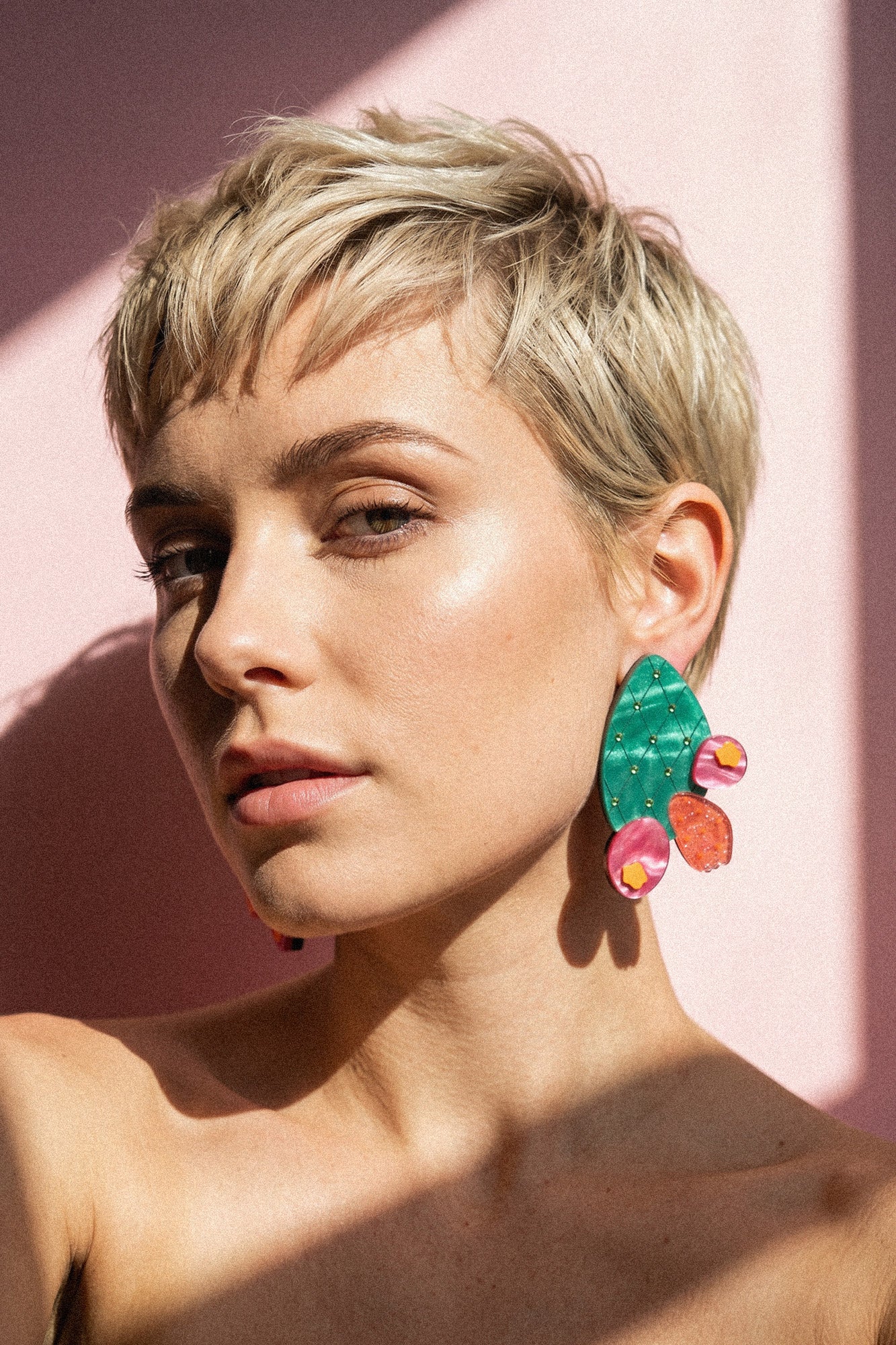 A woman with short blonde hair and glowy skin poses in sunlight, wearing bold design statement earrings shaped like opuncia cactus with green, pink, and orange details, against a pink background.