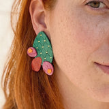 Close-up of a person with red hair wearing bold, statement earrings shaped like an opuncia cactus with pink and red flower accents. Only half of the face is visible, highlighting the earring's vibrant design and freckles.
