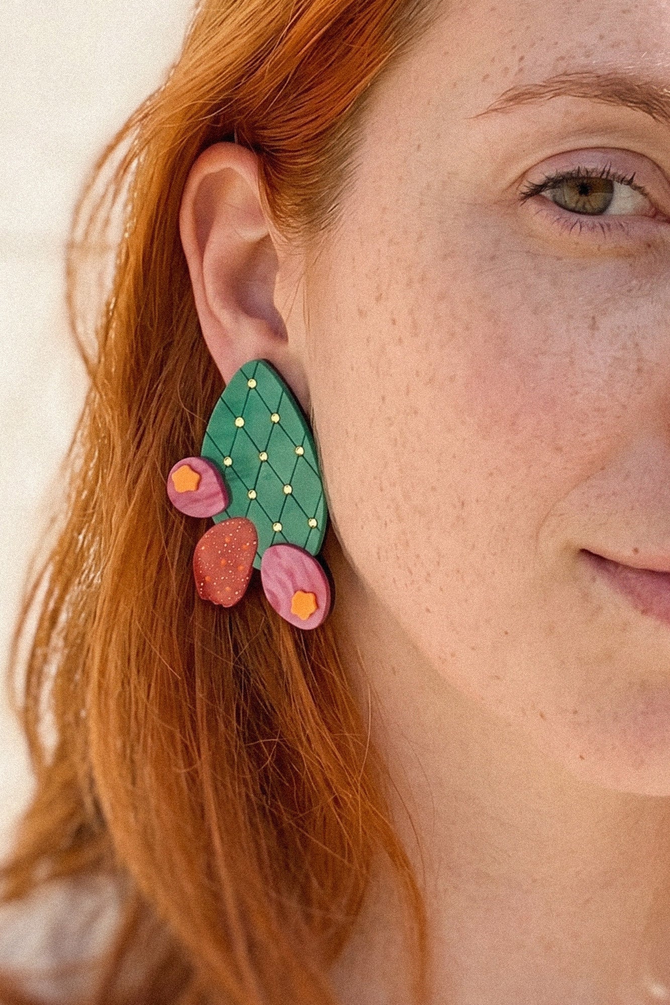 Close-up of a person with red hair wearing bold, statement earrings shaped like an opuncia cactus with pink and red flower accents. Only half of the face is visible, highlighting the earring's vibrant design and freckles.