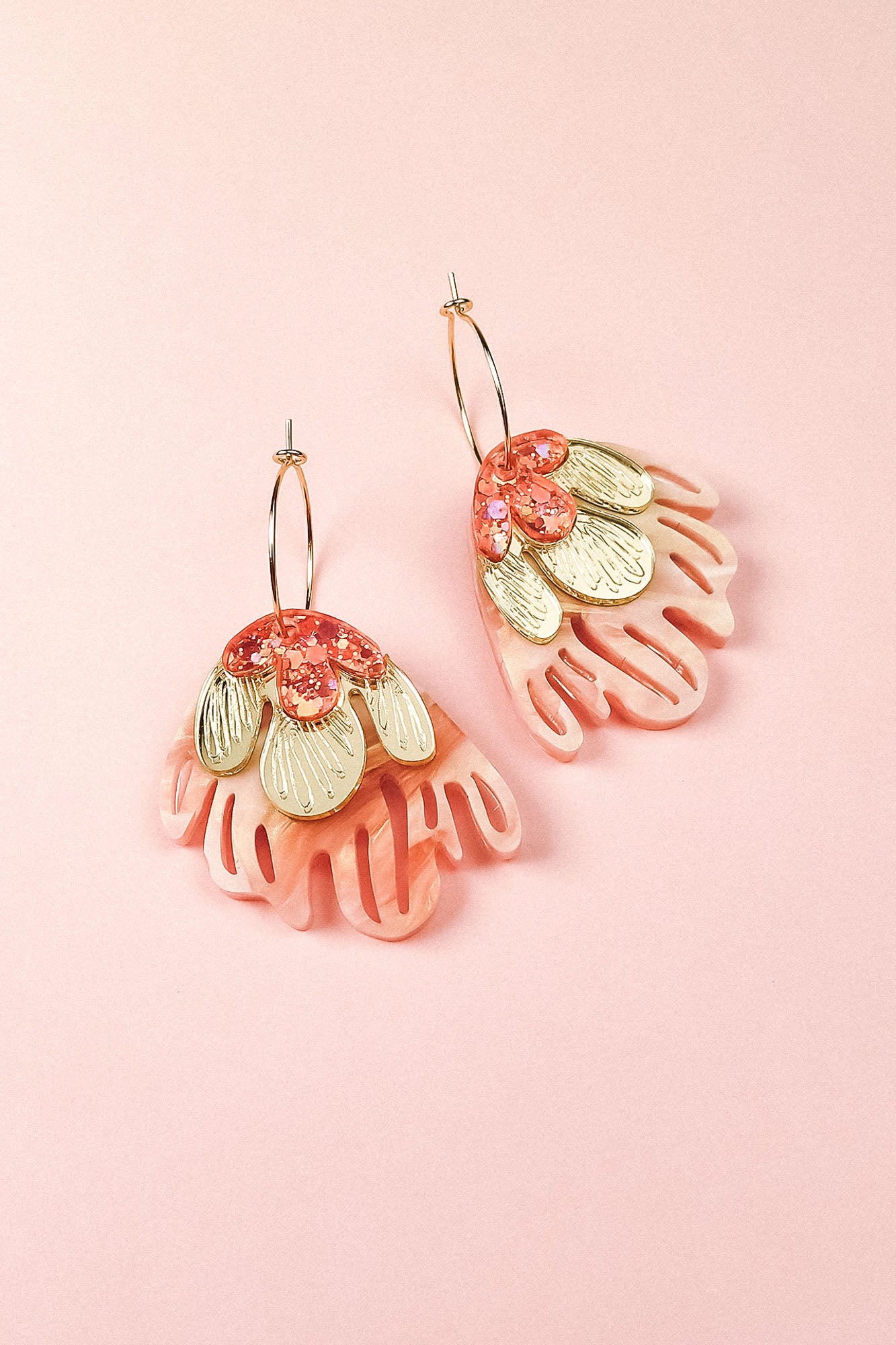 A pair of gold hoops with layered pink acrylic petals, gold-toned metal accents, and pink sequins evoke the charm of Organic Floral Hoops, beautifully arranged on a light pink background.