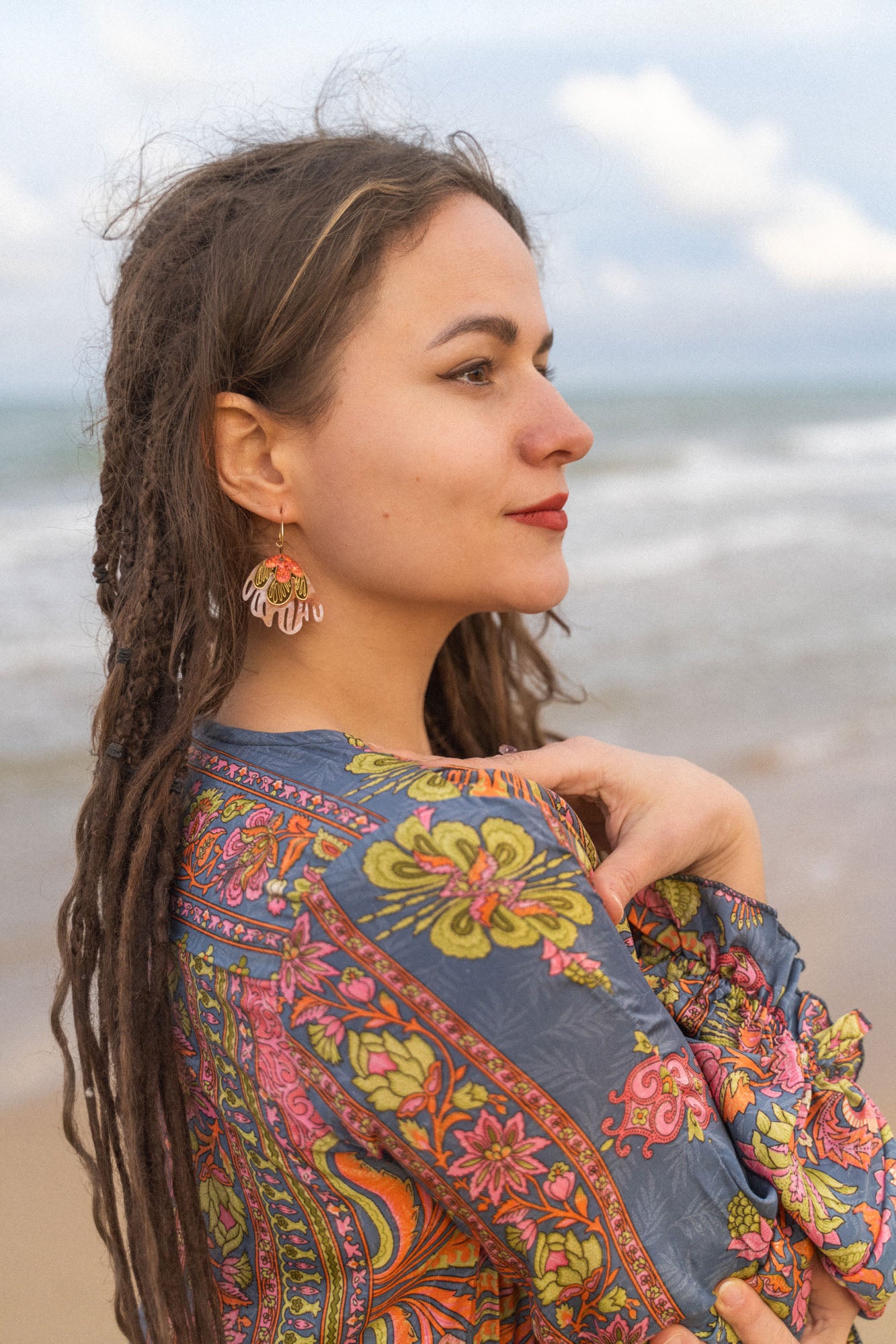 A woman with long, brown dreadlocks stands by the ocean, wearing a colorful, patterned shawl and Organic Floral Hoops accented with acrylic flowers, looking thoughtfully into the distance beneath a partly cloudy sky.