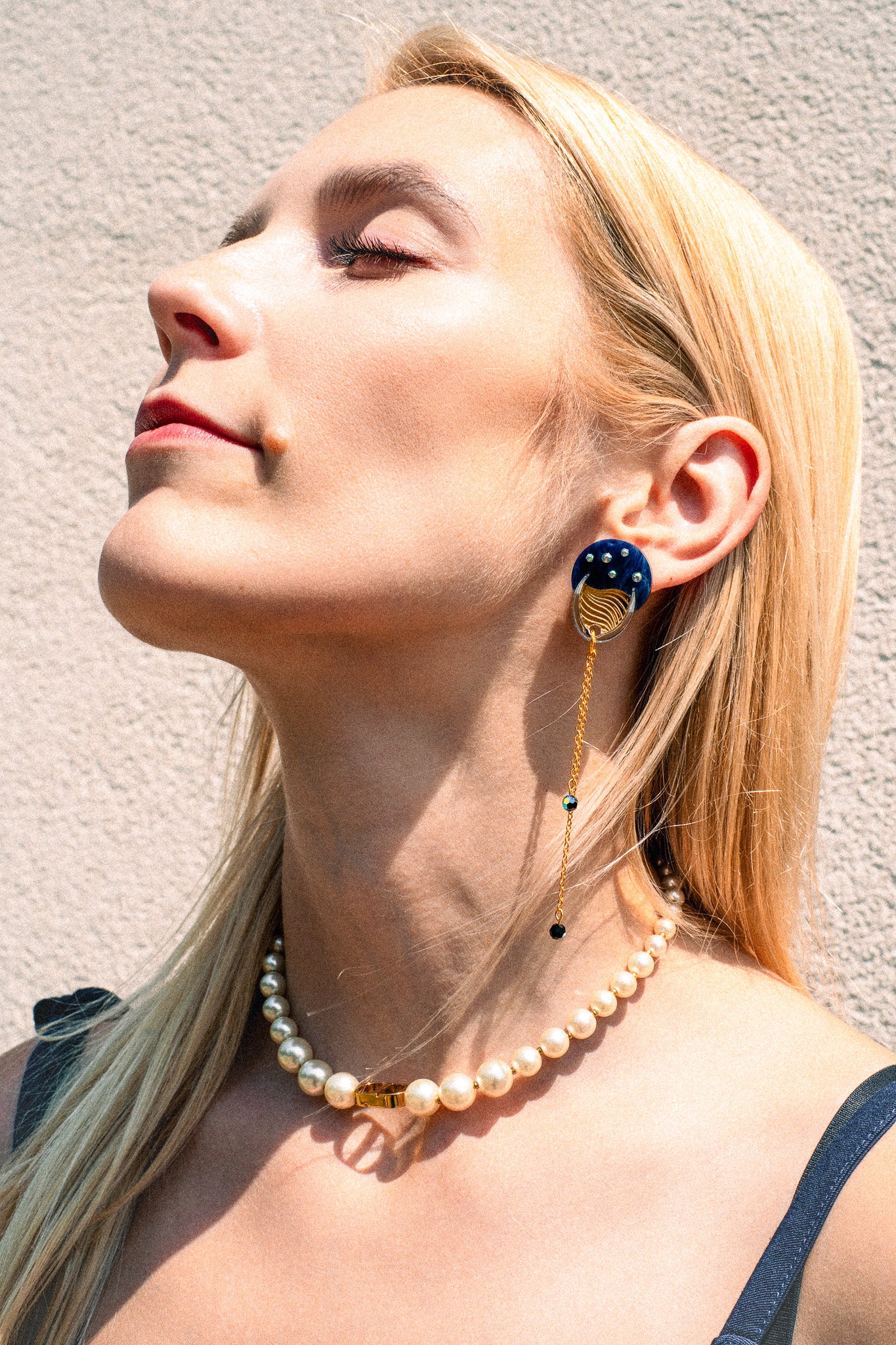 A woman with long blonde hair wears a pearl necklace and a bold blue and gold earring with chain detail, evoking festival magic as she poses in sunlight against a textured light wall.