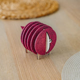 A set of round, maroon felt coasters stacked vertically on a metal holder shaped like a sheep, placed on a light wooden table near woven placemats—an ideal housewarming gift.