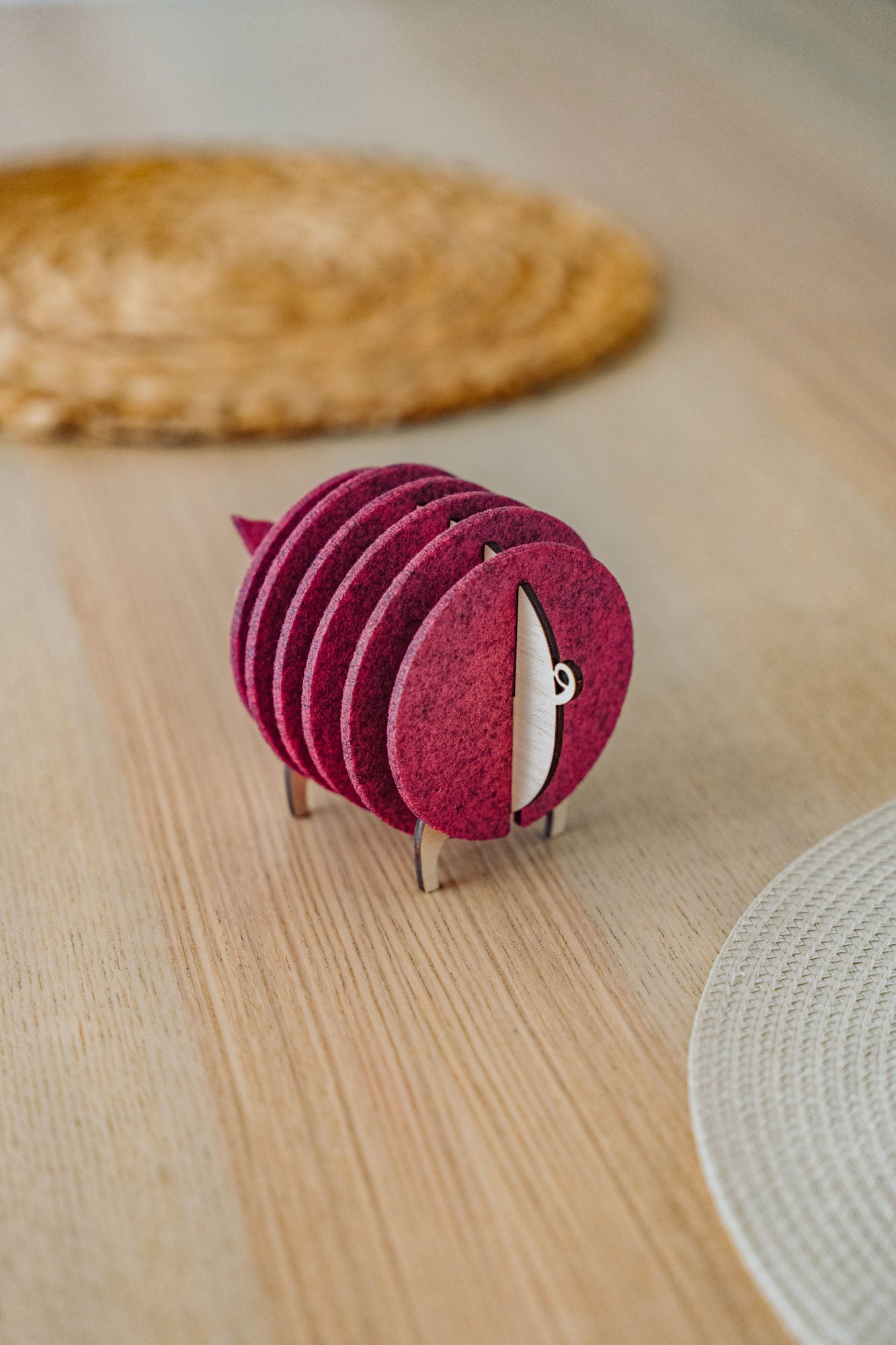 A set of round, maroon felt coasters stacked vertically on a metal holder shaped like a sheep, placed on a light wooden table near woven placemats—an ideal housewarming gift.