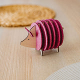 A wooden and felt coaster holder shaped like a hedgehog, with pink felt circles as its body, sits on a light wooden table near a woven placemat—an adorable housewarming gift for any home.