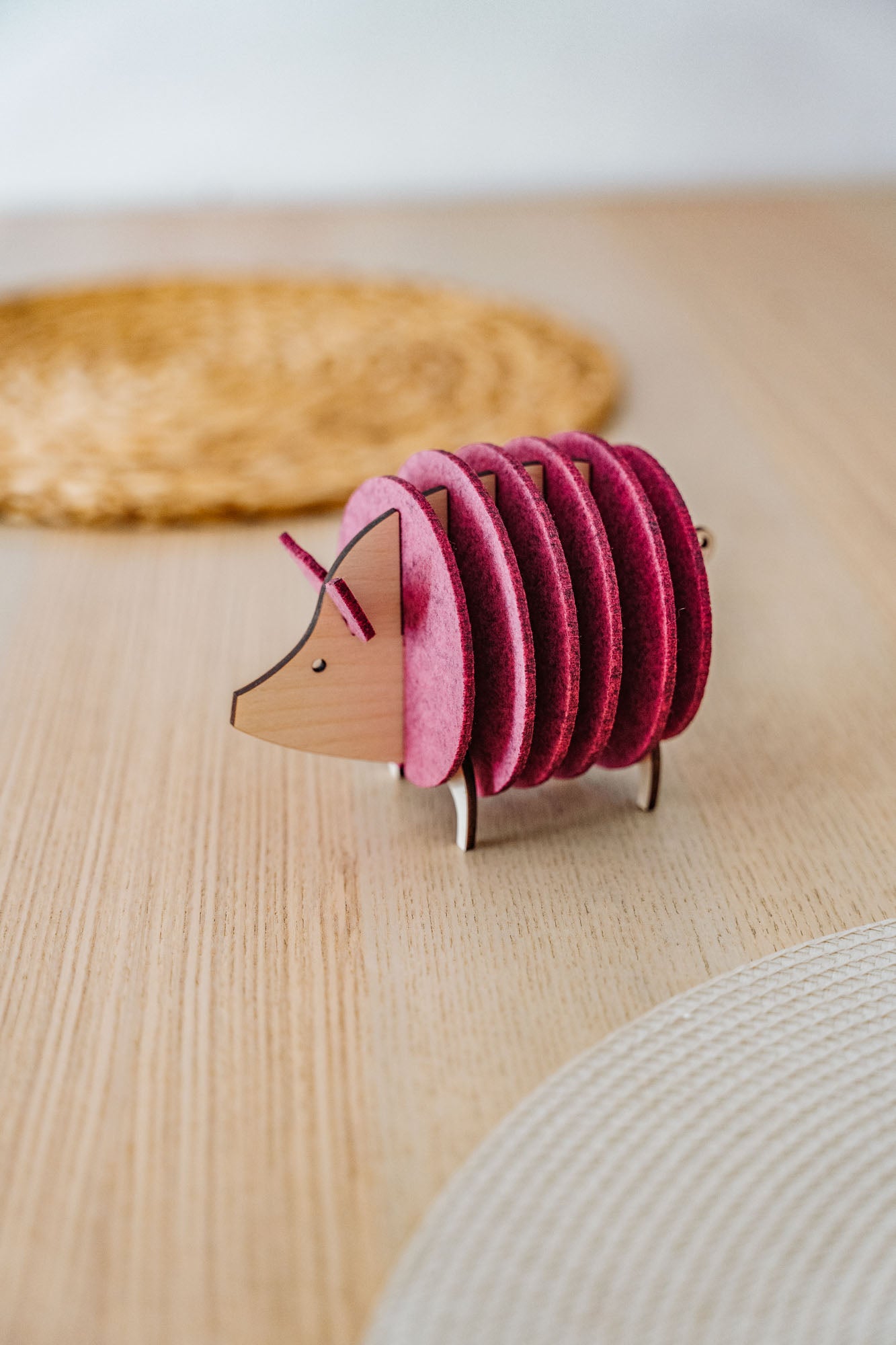 A wooden and felt coaster holder shaped like a hedgehog, with pink felt circles as its body, sits on a light wooden table near a woven placemat—an adorable housewarming gift for any home.