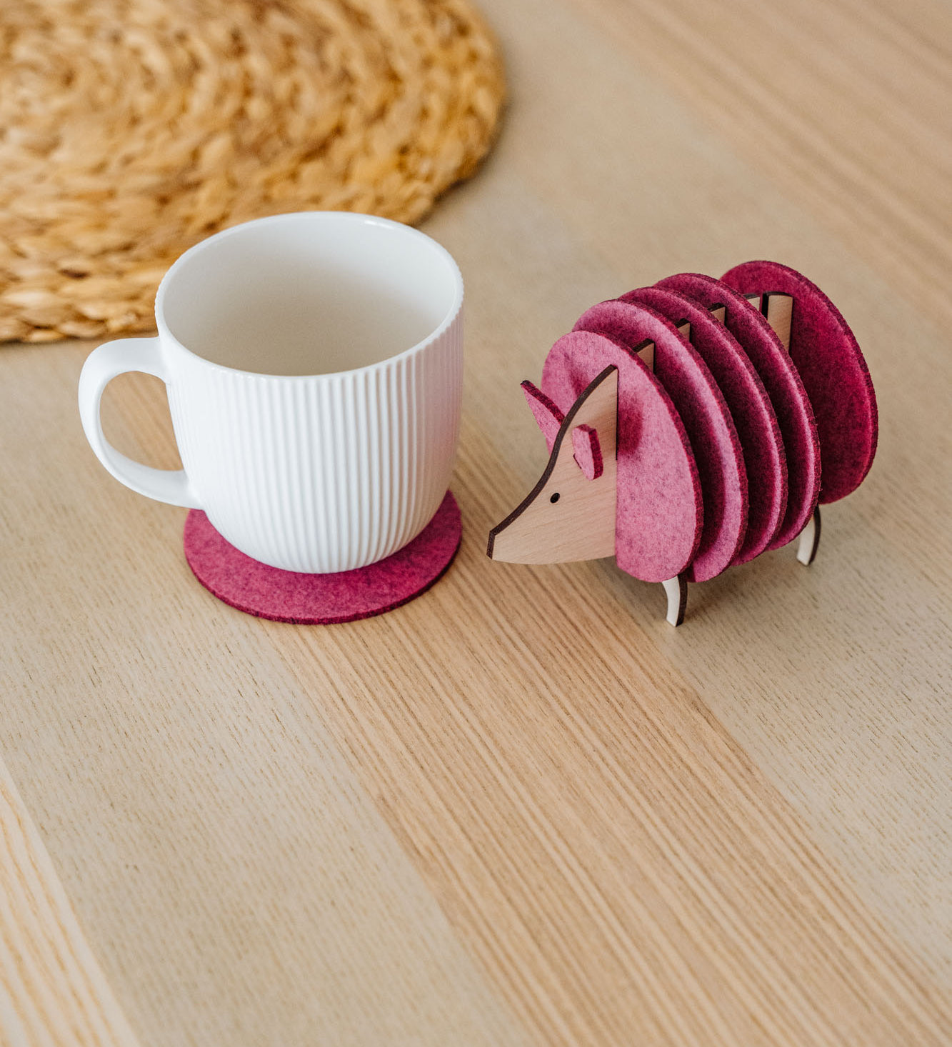 A white ribbed mug on a pink felt coaster sits next to a pink, hedgehog-shaped coaster holder on a light wooden table. A woven placemat is partially visible—perfect as a charming housewarming gift.