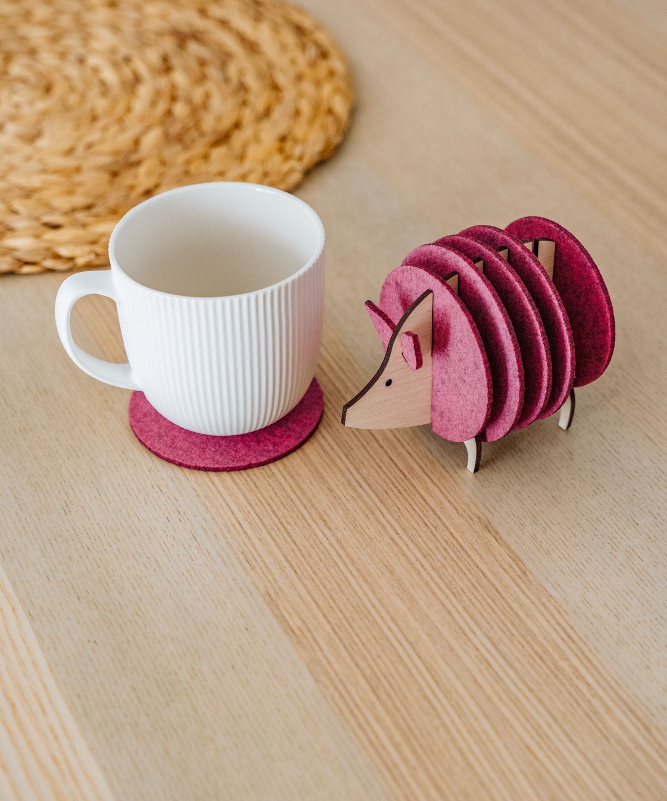 A white ribbed mug on a pink felt coaster sits next to a pink, hedgehog-shaped coaster holder on a light wooden table. A woven placemat is partially visible—perfect as a charming housewarming gift.