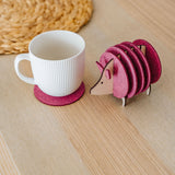 A white ribbed mug on a pink felt coaster sits next to a pink, hedgehog-shaped coaster holder on a light wooden table. A woven placemat is partially visible—perfect as a charming housewarming gift.