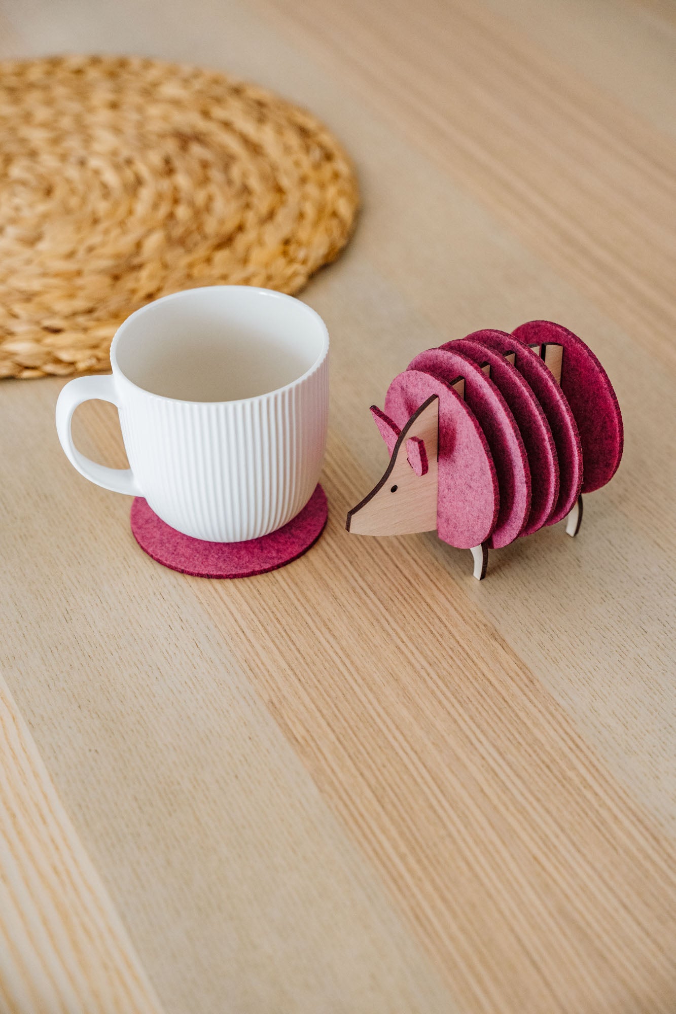 A white ribbed mug on a pink felt coaster sits next to a pink, hedgehog-shaped coaster holder on a light wooden table. A woven placemat is partially visible—perfect as a charming housewarming gift.