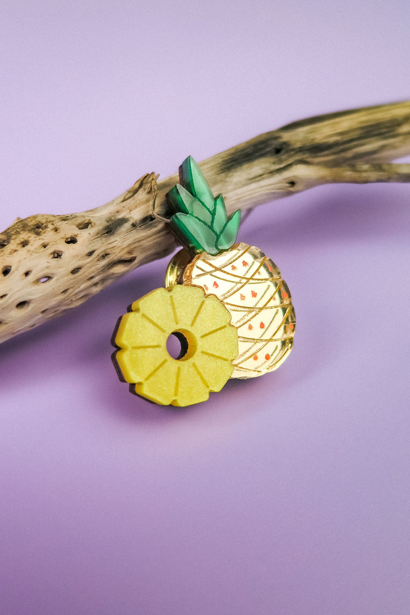 A decorative handcrafted Pineapple Pin with green leaves and a sliced section rests on a piece of driftwood against a pastel purple background.
