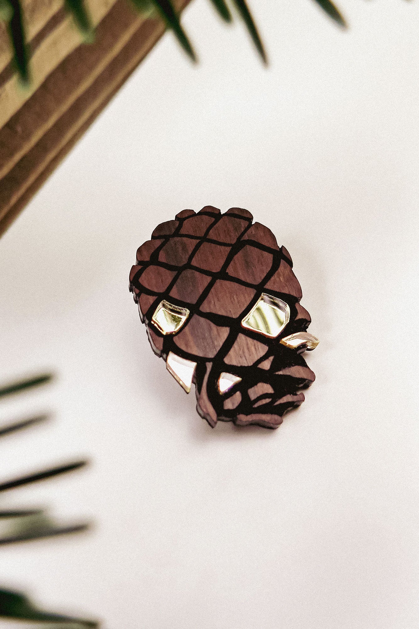 A brown, pinecone-shaped brooch crafted from recycled wood with shiny accents sits on a white surface, surrounded by blurred green leaves and a wooden object in the background.