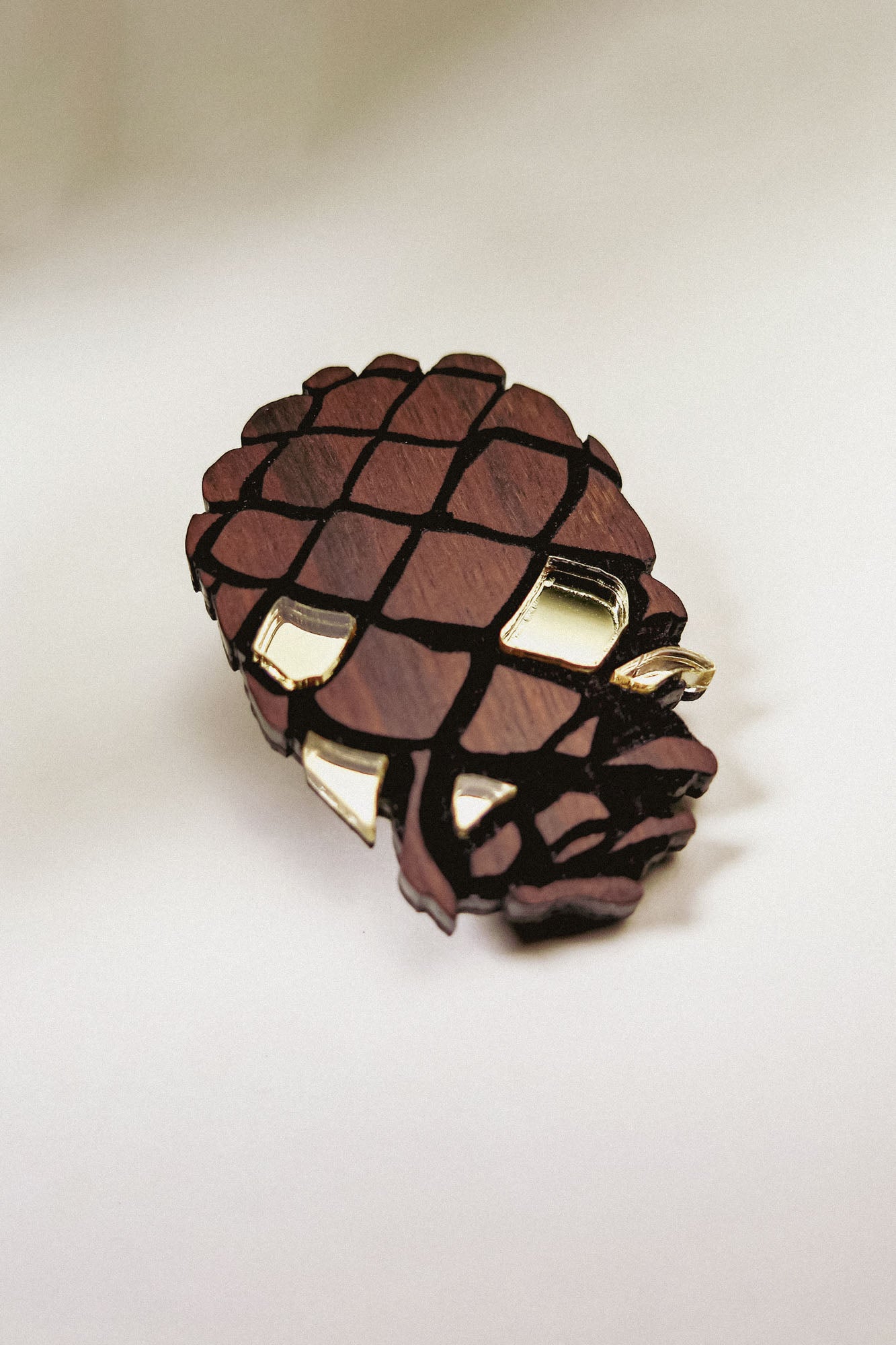 A sustainable Pinecone Brooch, crafted from recycled wood with gold accents, sits on a white surface, casting a soft shadow.