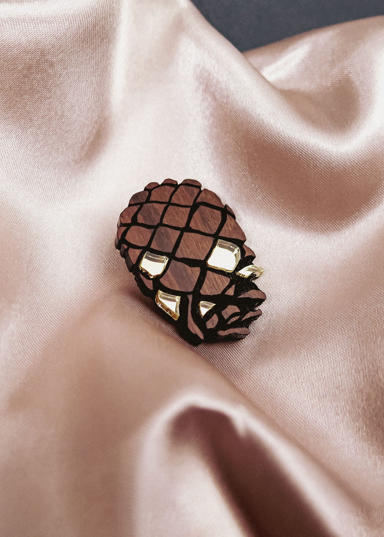 A gold and brown Pinecone Brooch, crafted from sustainable recycled wood, rests on shiny, light beige satin fabric with soft folds.