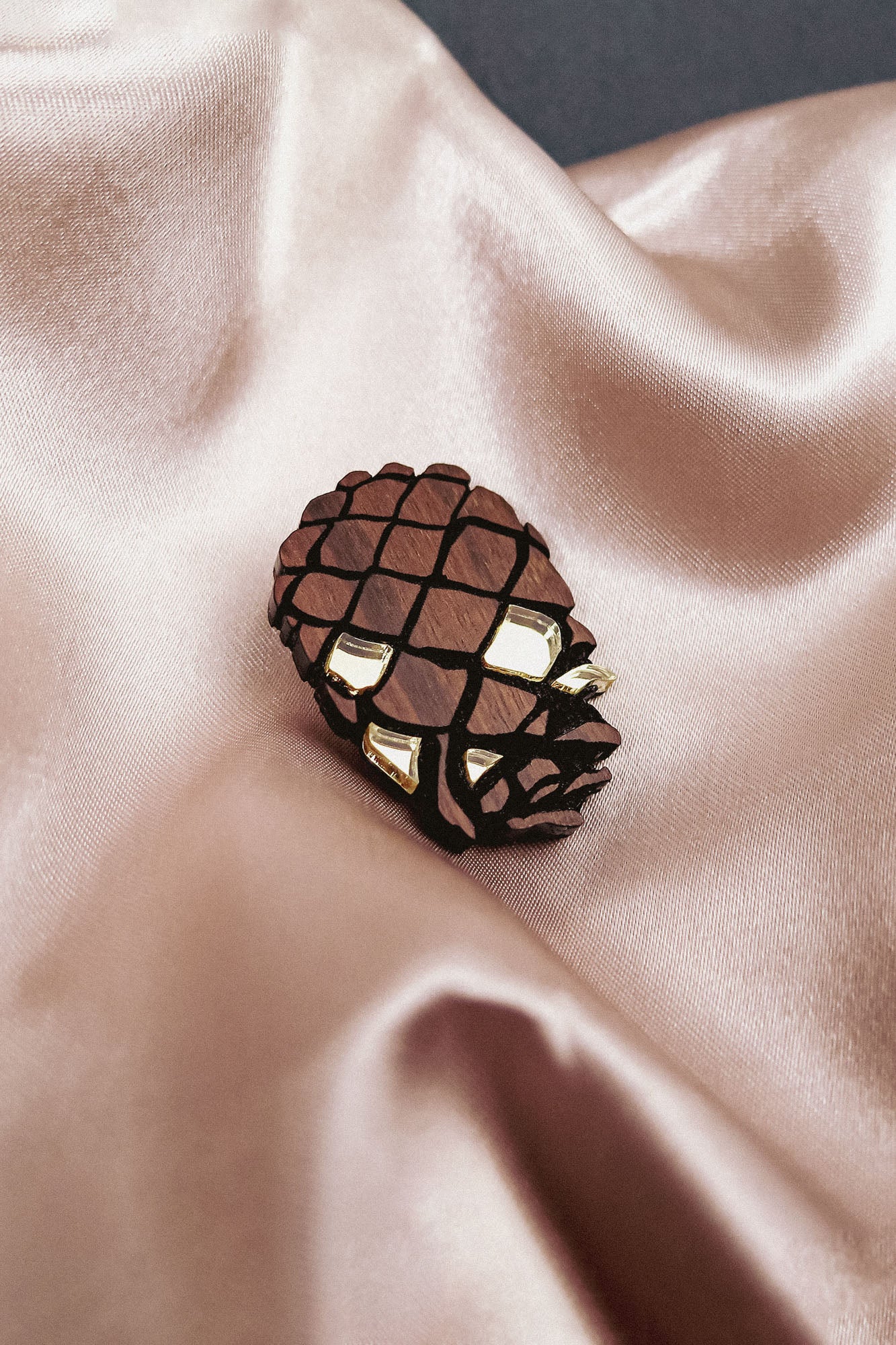 A gold and brown Pinecone Brooch, crafted from sustainable recycled wood, rests on shiny, light beige satin fabric with soft folds.
