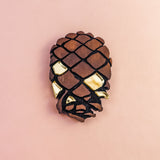 A Pinecone Brooch crafted from sustainable, recycled wood with gold accents is displayed against a light pink background, shaped like a pinecone or stylized skull.