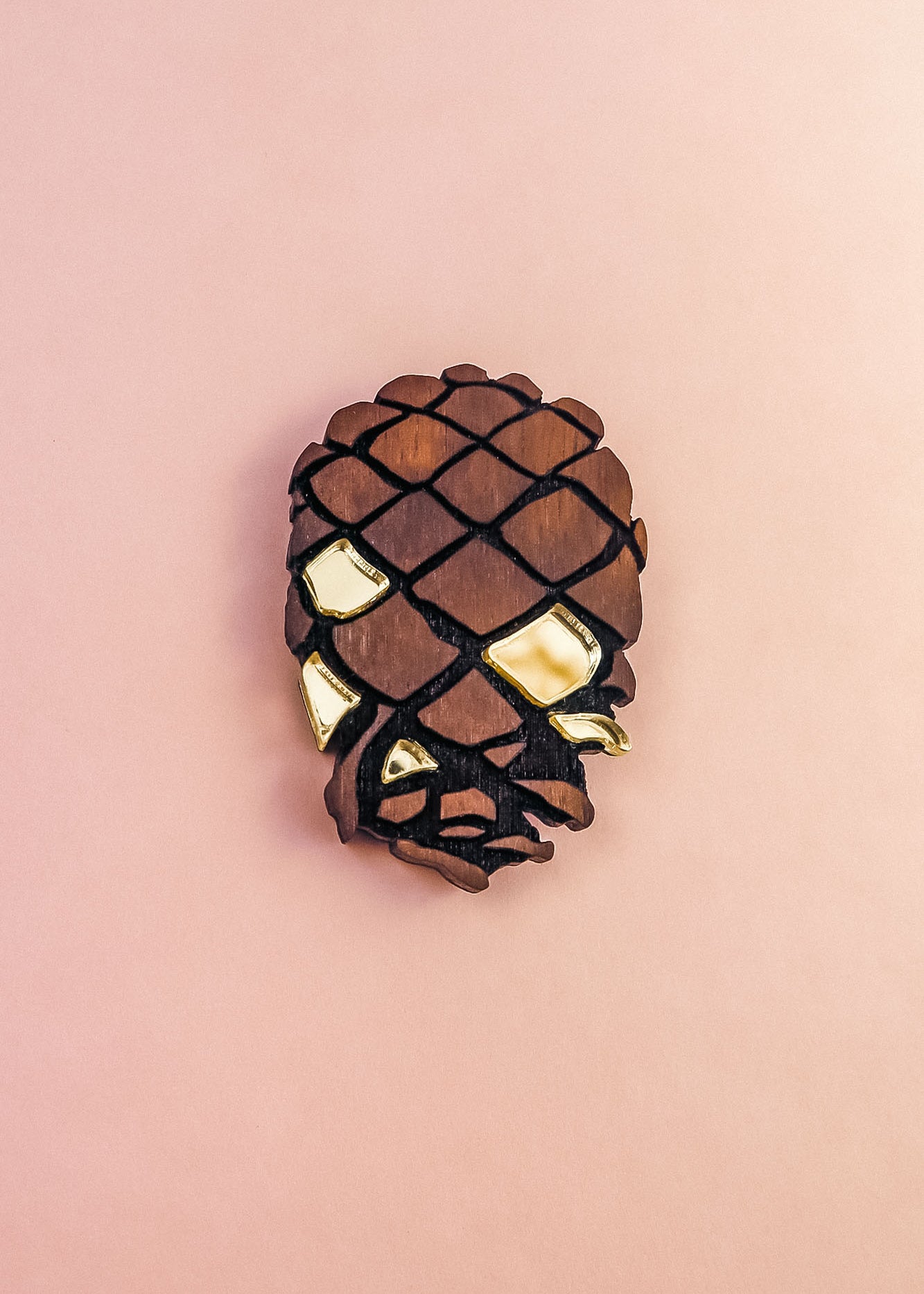 A Pinecone Brooch crafted from sustainable, recycled wood with gold accents is displayed against a light pink background, shaped like a pinecone or stylized skull.