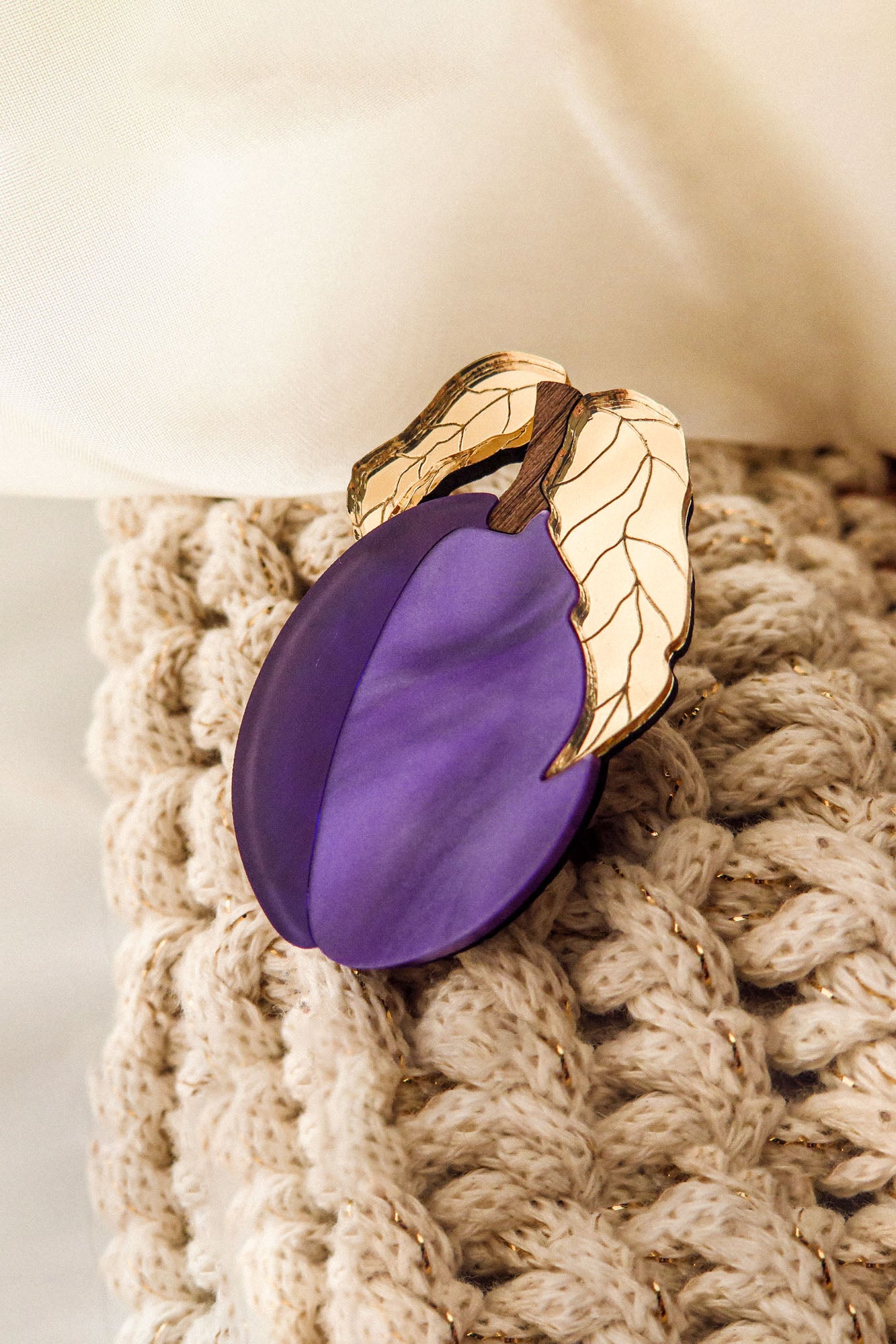 A handmade purple plum-shaped brooch with two gold and ivory leaves is attached to a chunky beige knit fabric. Cream-colored fabric appears in the background.