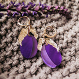 Pair of gold and purple plum earrings with textured gold accents, displayed on a knitted beige fabric with a sprig of purple flowers alongside. These handcrafted earrings add a touch of elegance to any outfit.