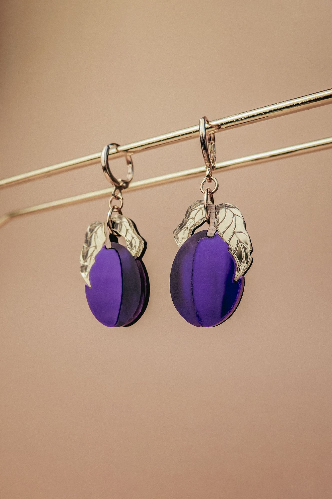 A pair of elegant purple plum earrings with silver leaf accents hang from a gold-colored rod against a beige background.