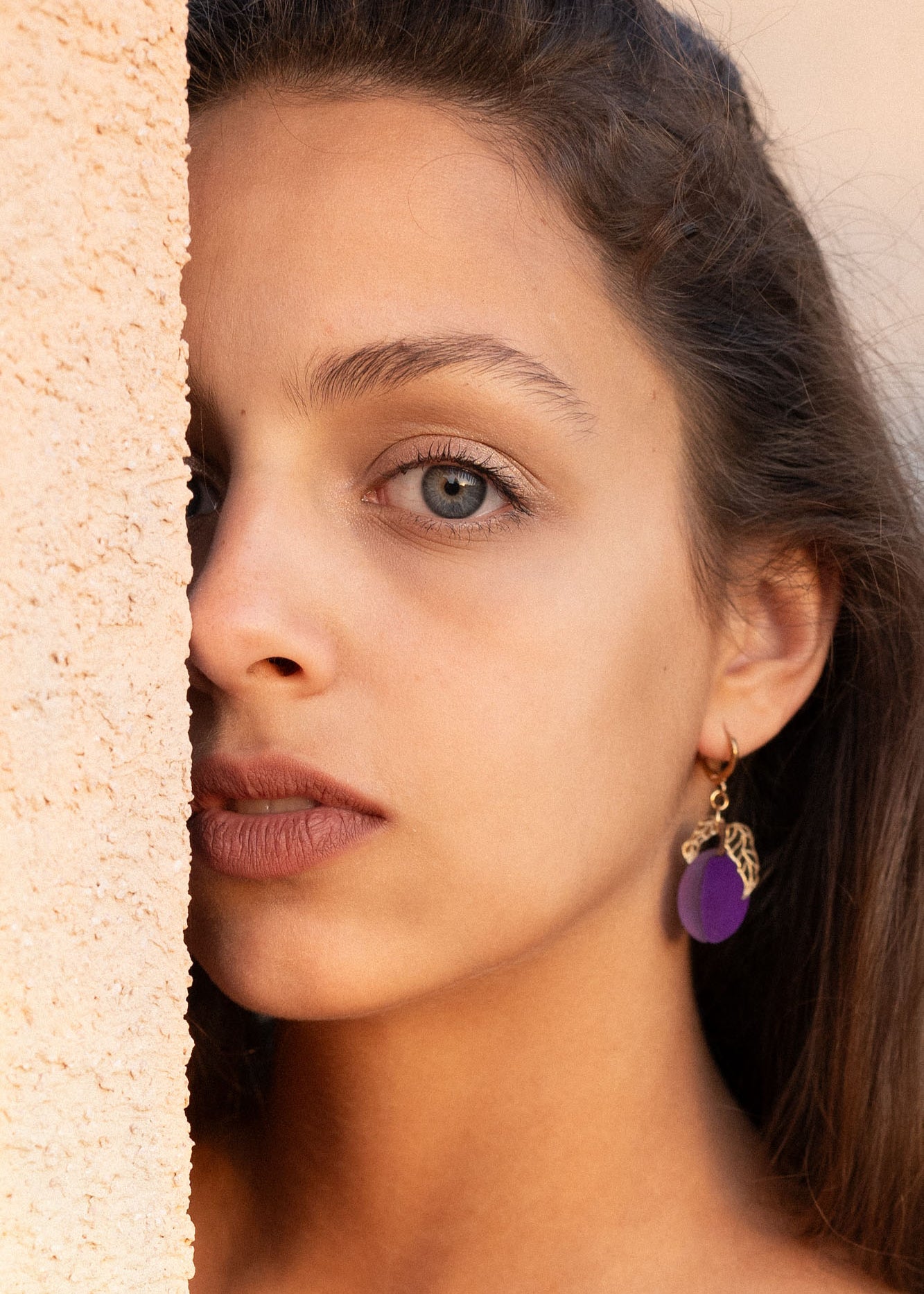 A woman with long brown hair and elegant purple plum earrings looks directly at the camera, standing next to a textured beige wall, with soft natural light highlighting her face.