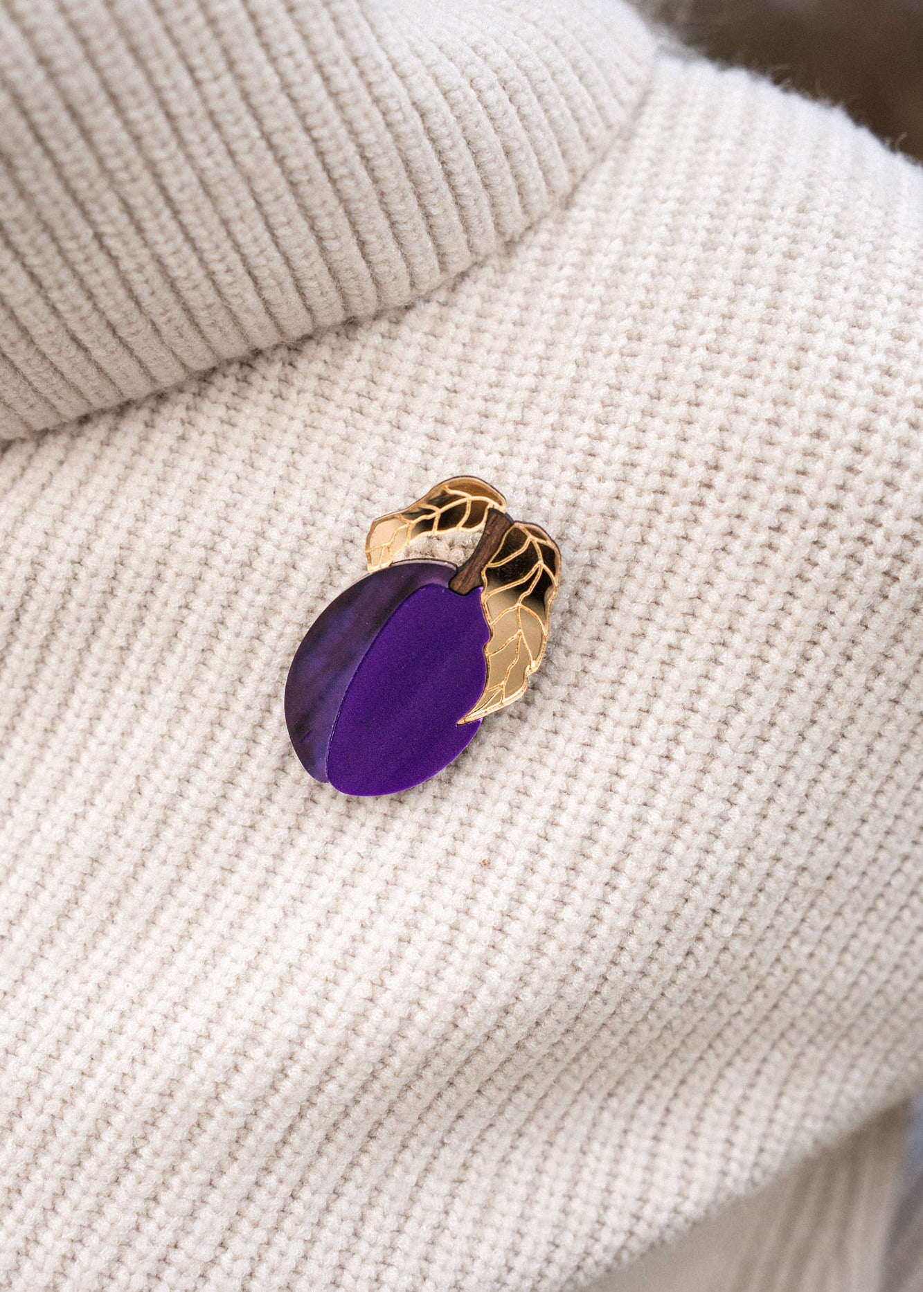 A handcrafted brooch shaped like a gold and purple eggplant with two gold leaves is pinned to a cream-colored ribbed sweater.