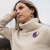 A woman with blonde hair wearing a cream-colored turtleneck sweater, handcrafted purple plum earrings, and a matching purple brooch stands outdoors, looking thoughtful with her hand resting on her head.