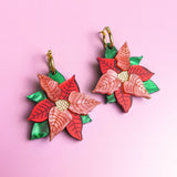 A pair of Poinsettia Statement Hoops shaped like poinsettia flowers with red and pink petals, yellow centers, and green leaves, displayed on a light purple background—perfect festive earrings for your holiday jewelry collection.