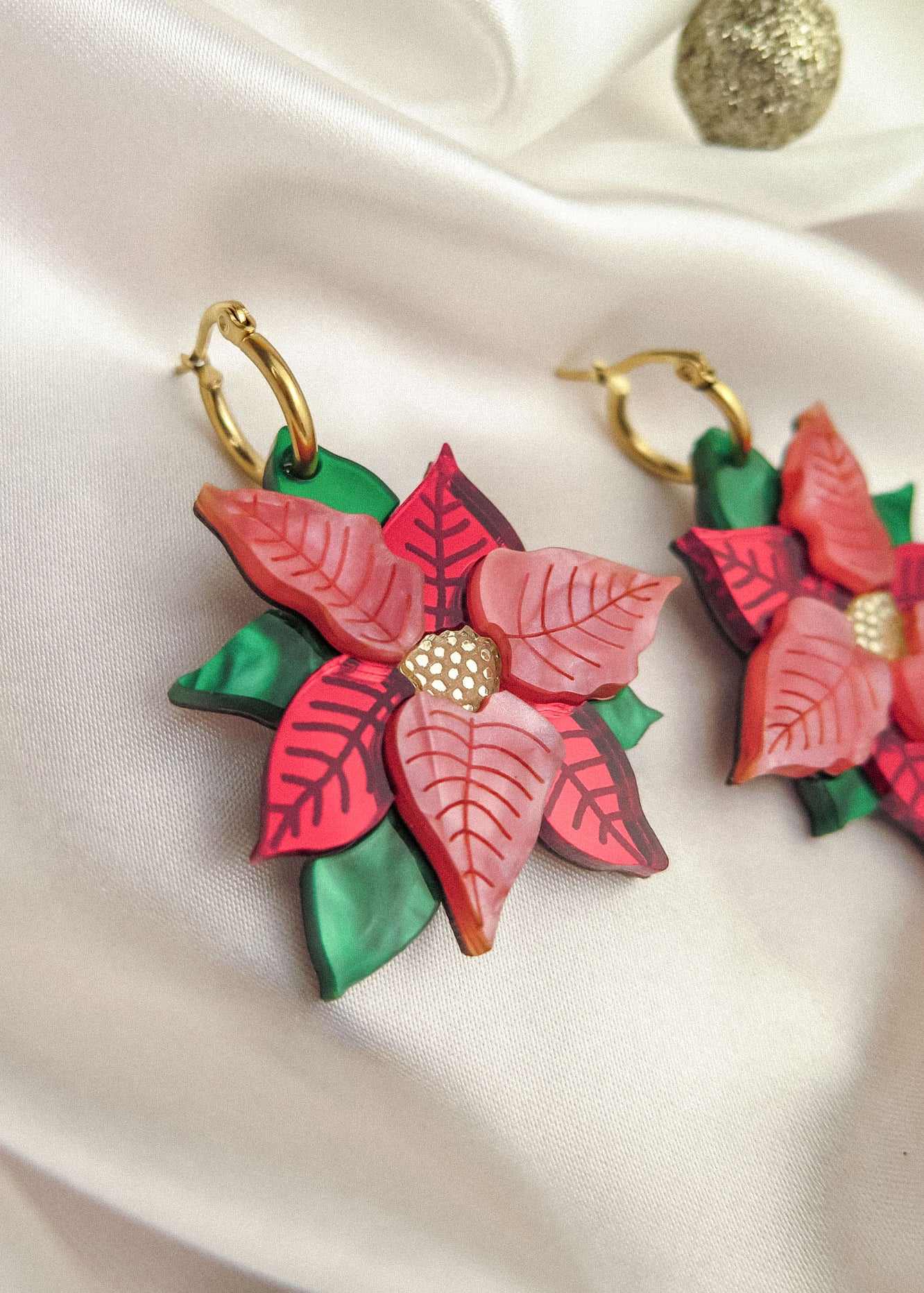Close-up of festive earrings called Poinsettia Statement Hoops, featuring red and pink petals, green leaves, and gold hoop hooks, placed on white satin with a small gold ornament—perfect holiday jewelry for the season.