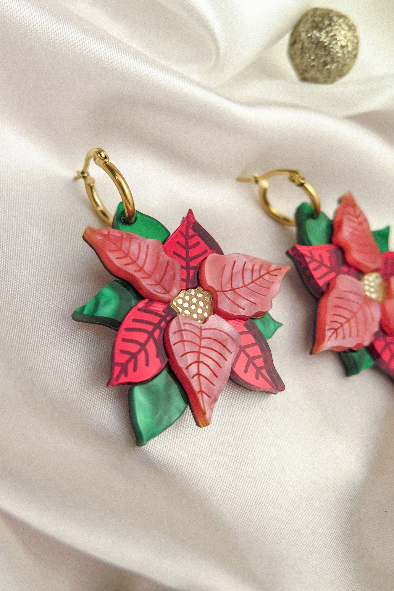 Close-up of festive earrings called Poinsettia Statement Hoops, featuring red and pink petals, green leaves, and gold hoop hooks, placed on white satin with a small gold ornament—perfect holiday jewelry for the season.