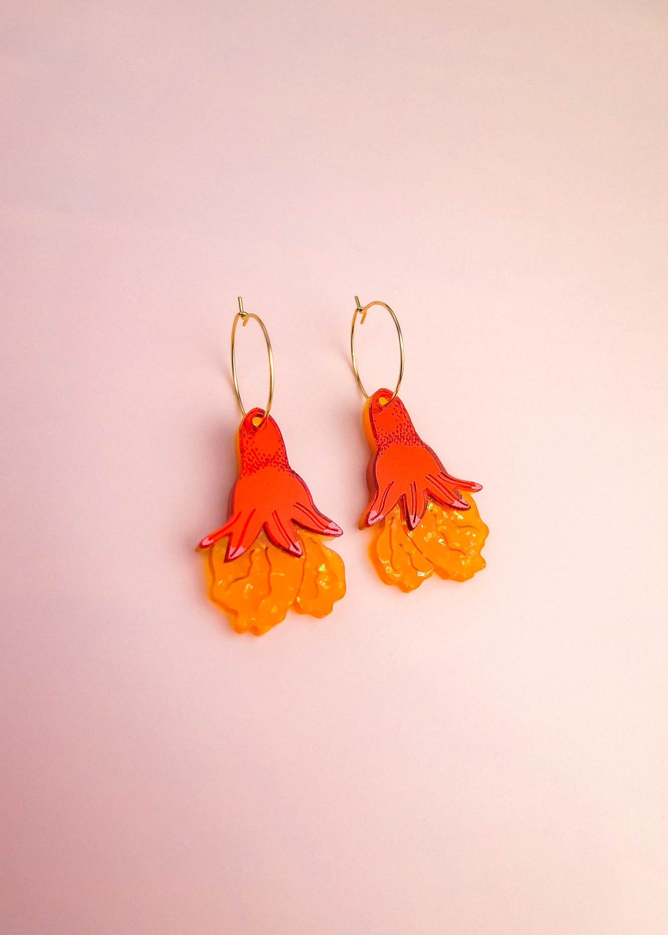 A pair of Little Turtle Hoop Earrings with gold hoops and dangling charms shaped like stylized orange and yellow flowers, displayed on a light pink background. These handcrafted earrings add a cheerful touch to any look.