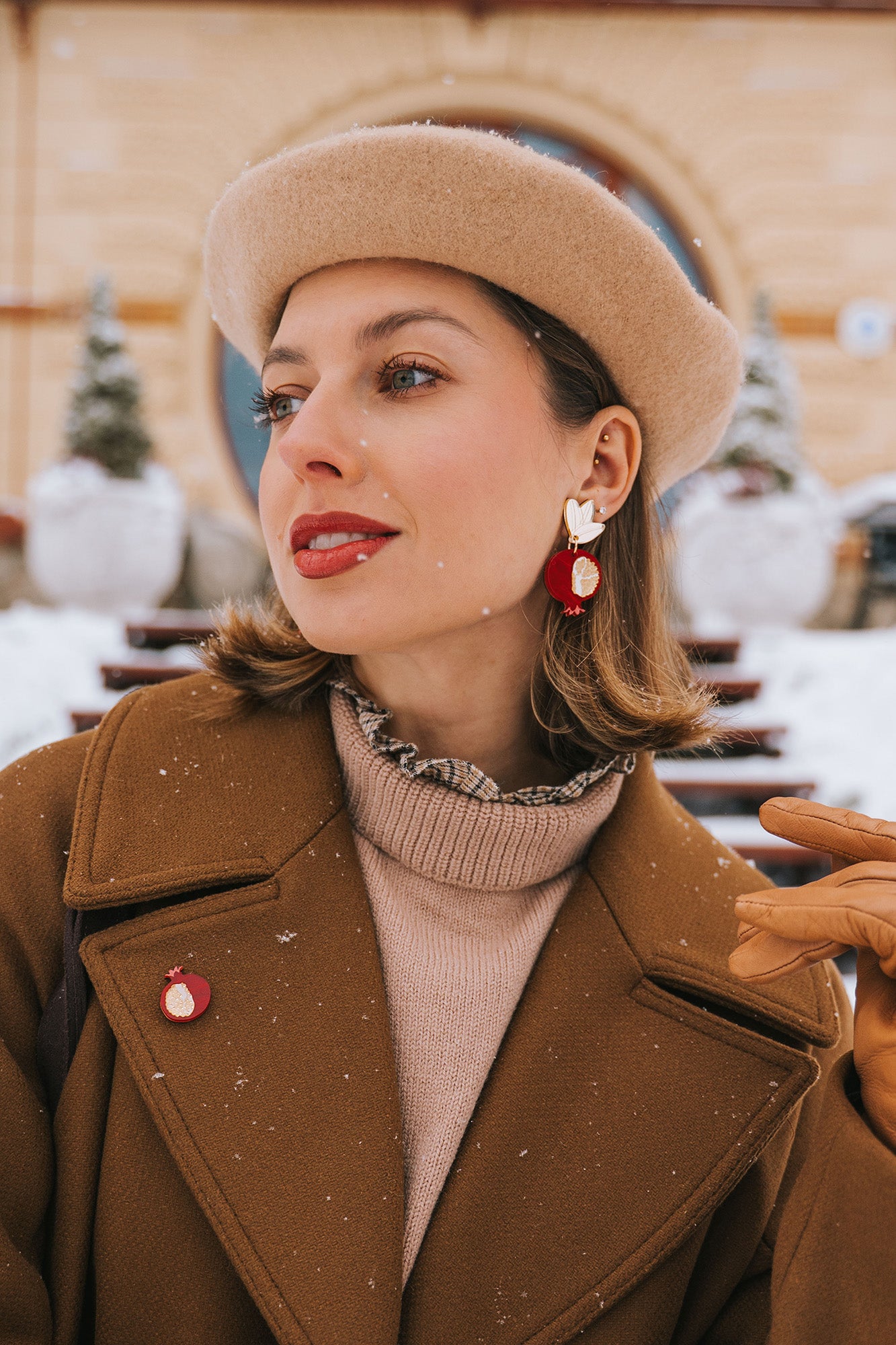 A woman in a tan beret and brown coat stands outside on a snowy day, wearing bold, handcrafted earrings with a bird design, a matching pin, tan gloves, and red lipstick. Snowflakes sprinkle her hair and coat.