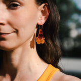 A woman with long brown hair wears handmade floral earrings and a sleeveless yellow top, standing outdoors in natural light. Only the left side of her face and shoulder are visible.