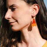 A woman with long brown hair wearing a sleeveless yellow top and bold handmade floral earrings stands outside in sunlight, smiling softly.