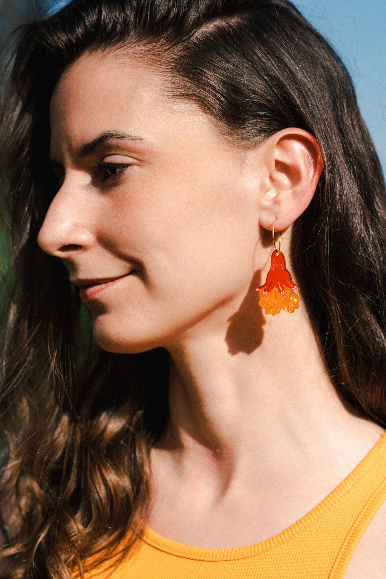 A woman with long brown hair wearing a sleeveless yellow top and bold handmade floral earrings stands outside in sunlight, smiling softly.
