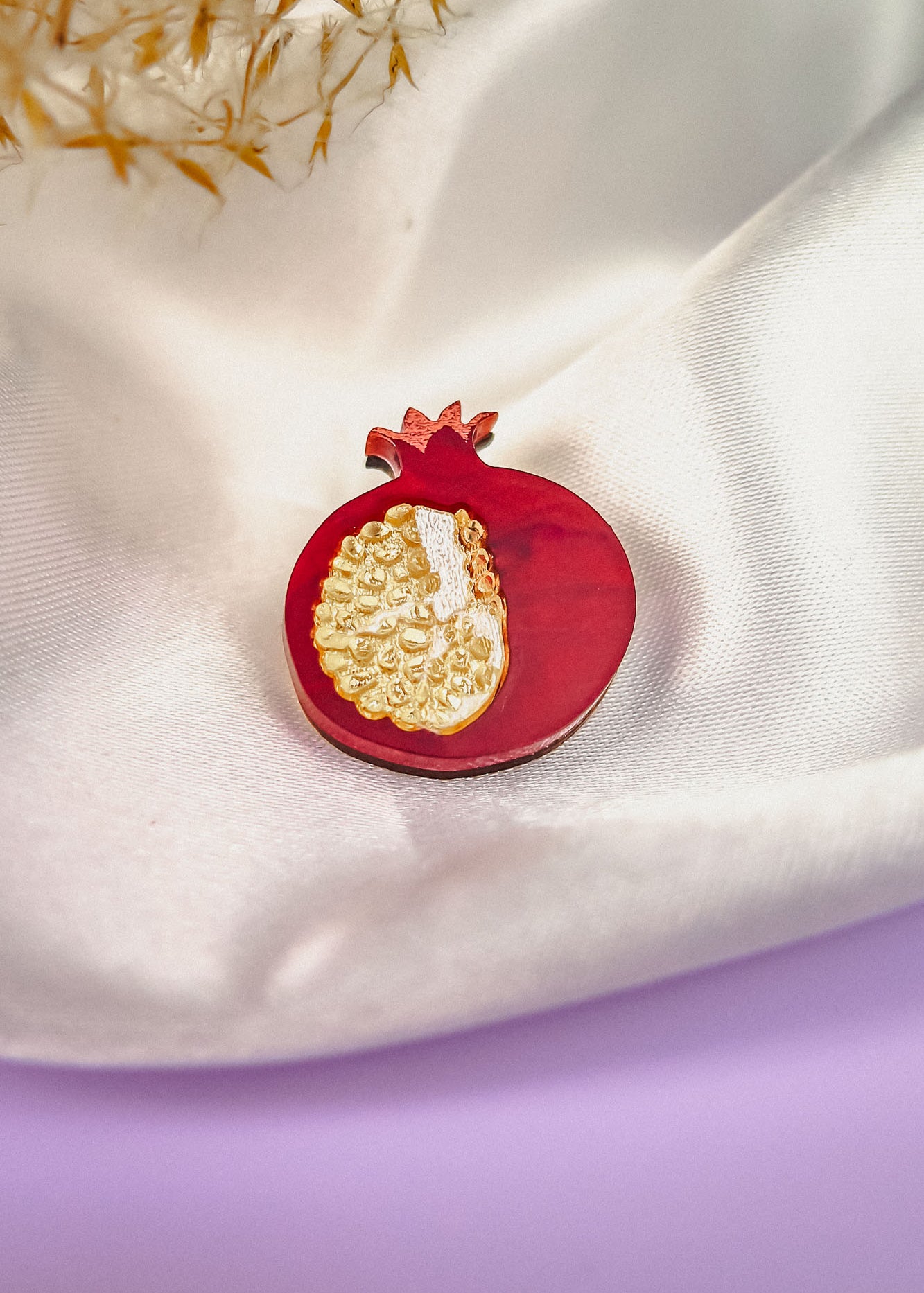 A handcrafted Pomegranate Pin, shaped like a red pomegranate with gold detailing, rests on white satin fabric. Some dried plants are visible in the background and a purple surface appears at the bottom—an elegant fashion accessory.