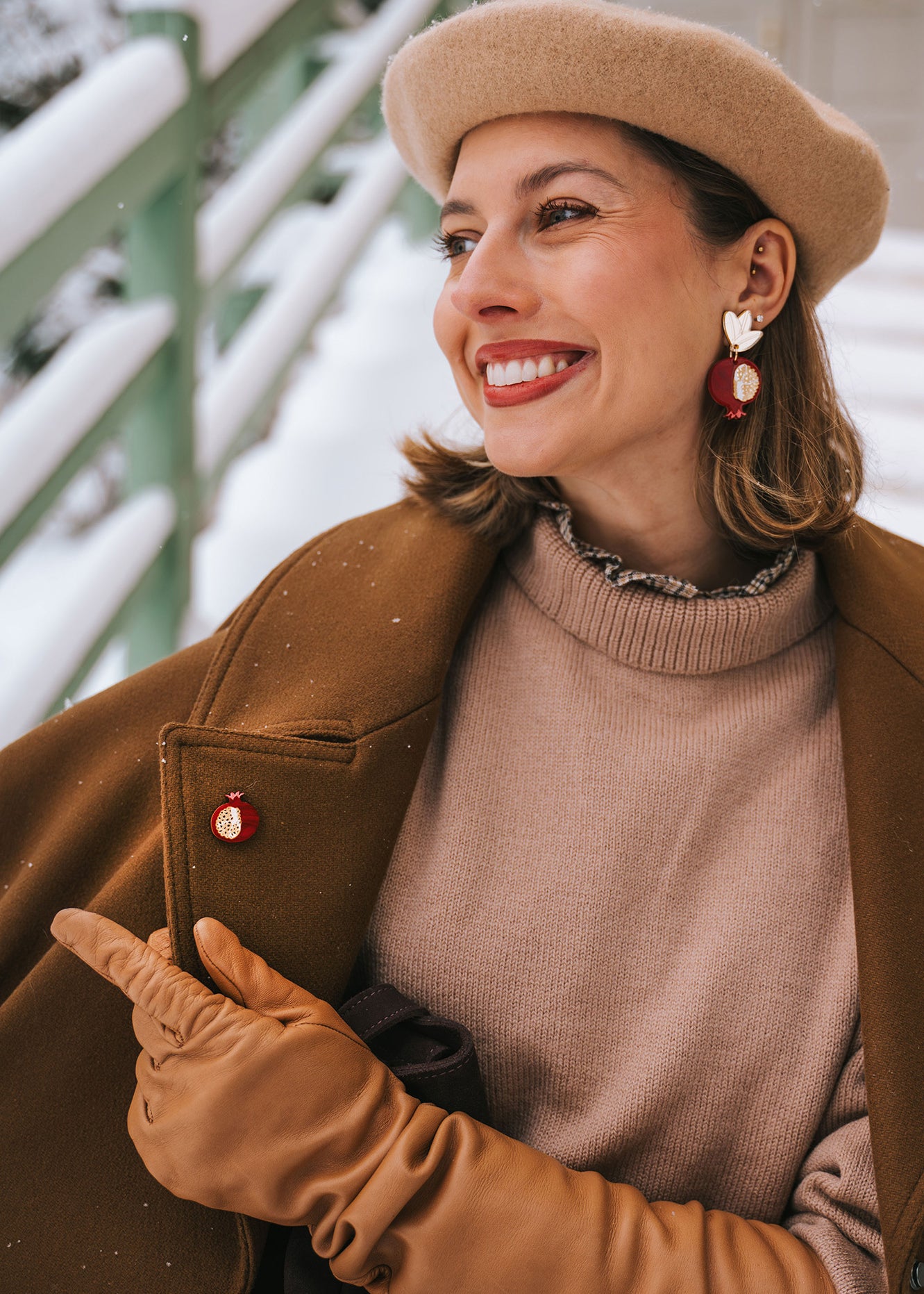 A smiling woman wearing a beige beret, brown coat, tan gloves, and statement earrings with red and gold accents sits outside in a snowy setting, pointing to a handcrafted Pomegranate Pin—a unique fashion accessory on her coat.
